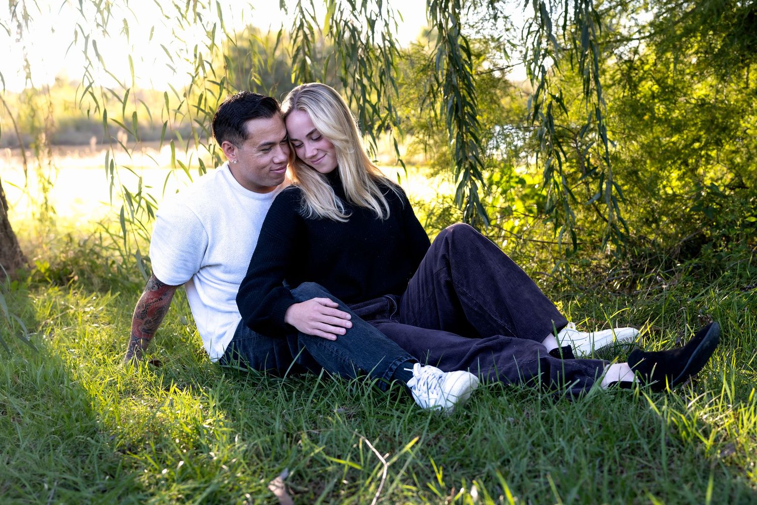 Smiling couple sitting close together on a grassy lakeside lawn under weeping willow branches on a sunny day.