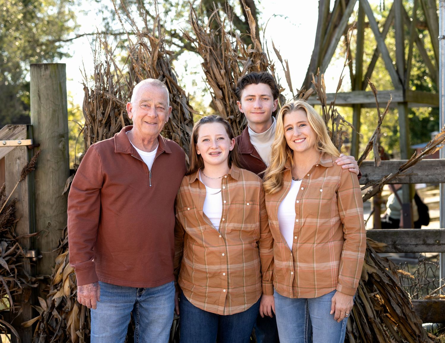 Four family members in matching rust and plaid shirts pose together outdoors in front of dried cornstalks and a rustic wooden fence.