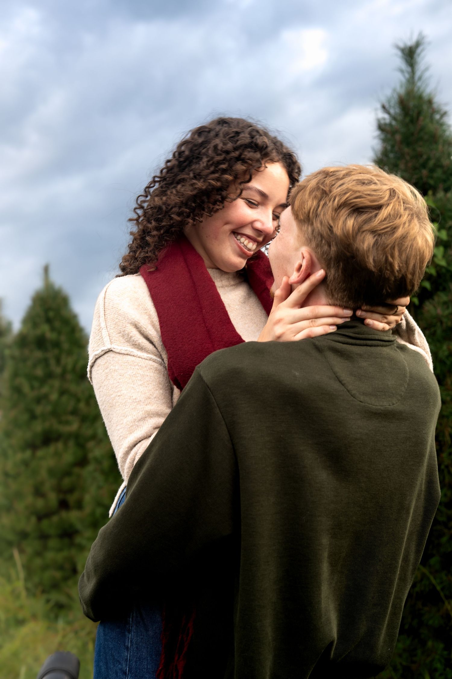 A joyful couple embraces outdoors near evergreen trees, sharing a romantic moment on a cloudy day.