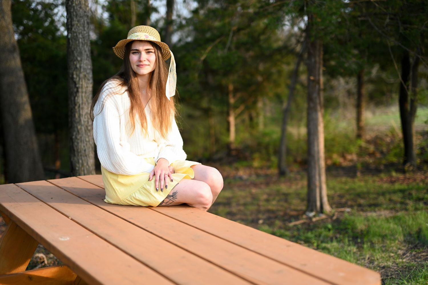 Young woman in straw hat and yellow skirt sitting cross-legged on a wooden bench outdoors.