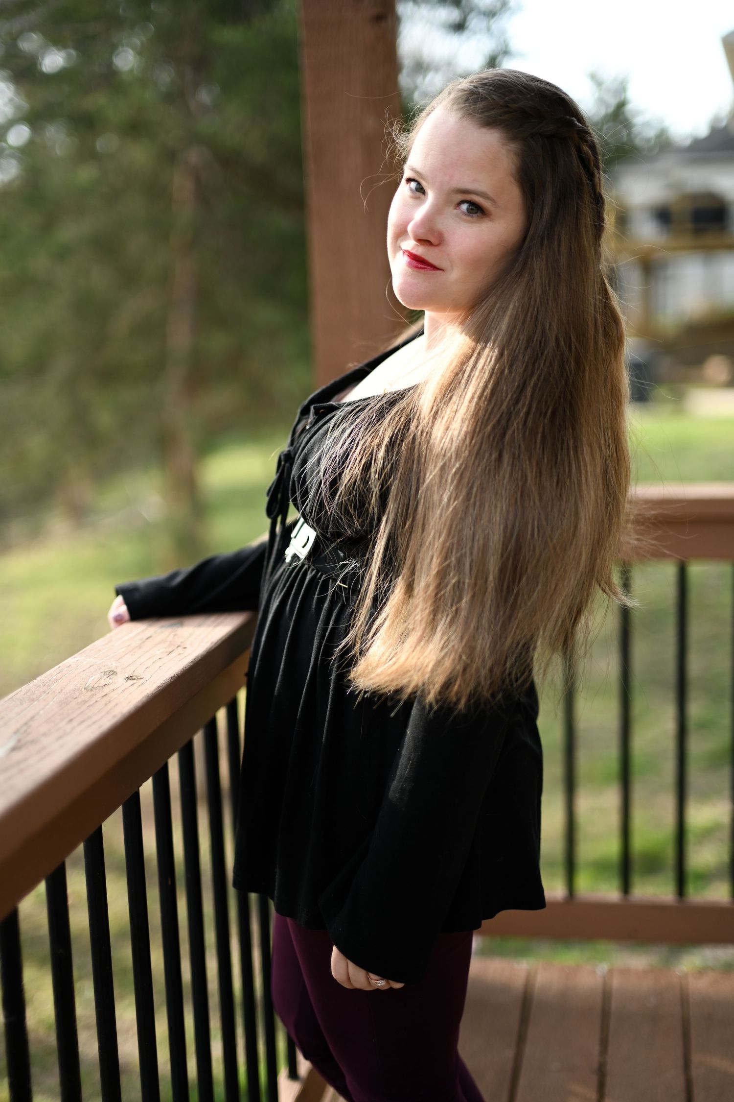 A young woman with long brown hair poses on a wooden deck railing wearing a black top and burgundy pants.