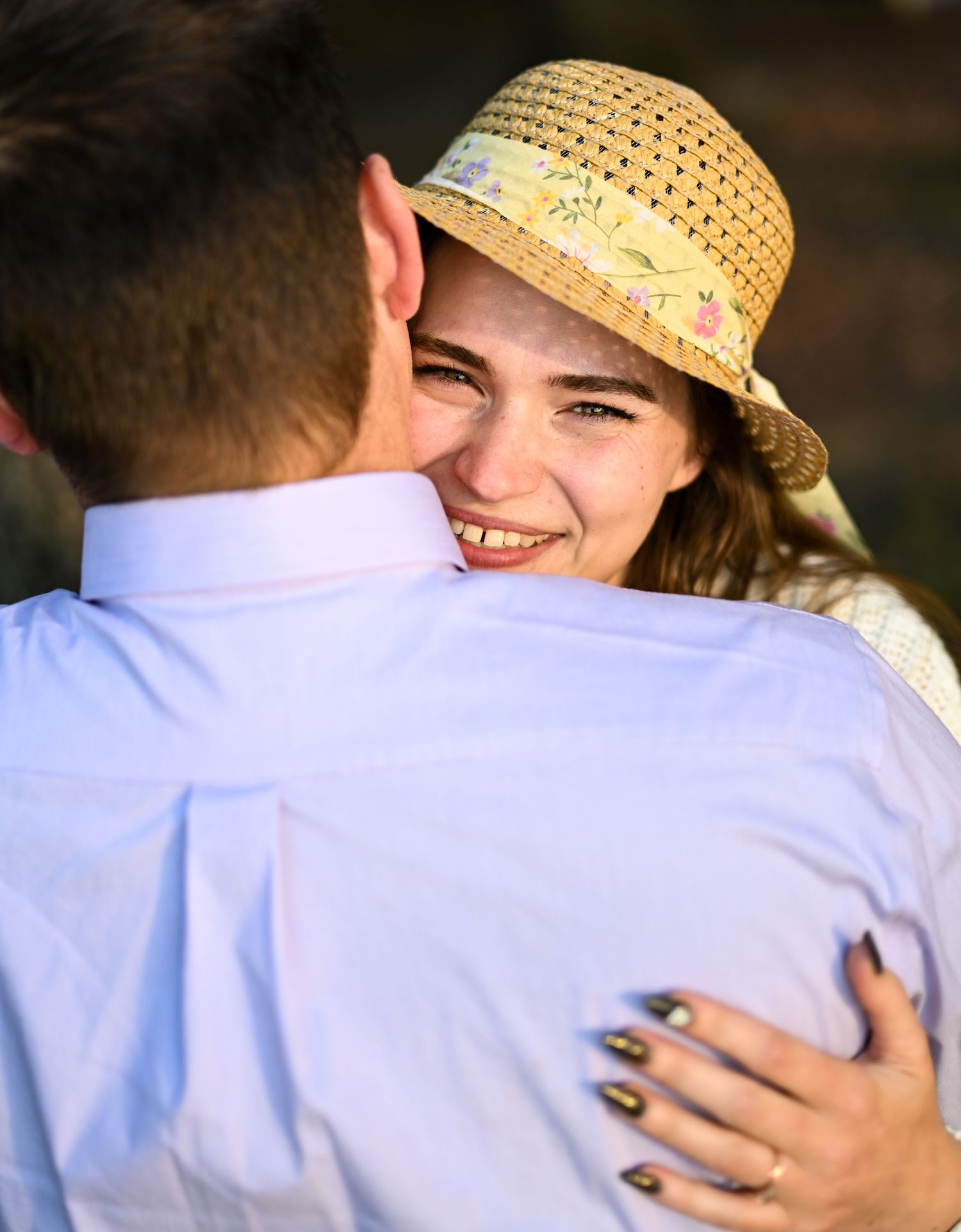 A joyful woman in a straw hat smiles while hugging a man in a light blue shirt outdoors.