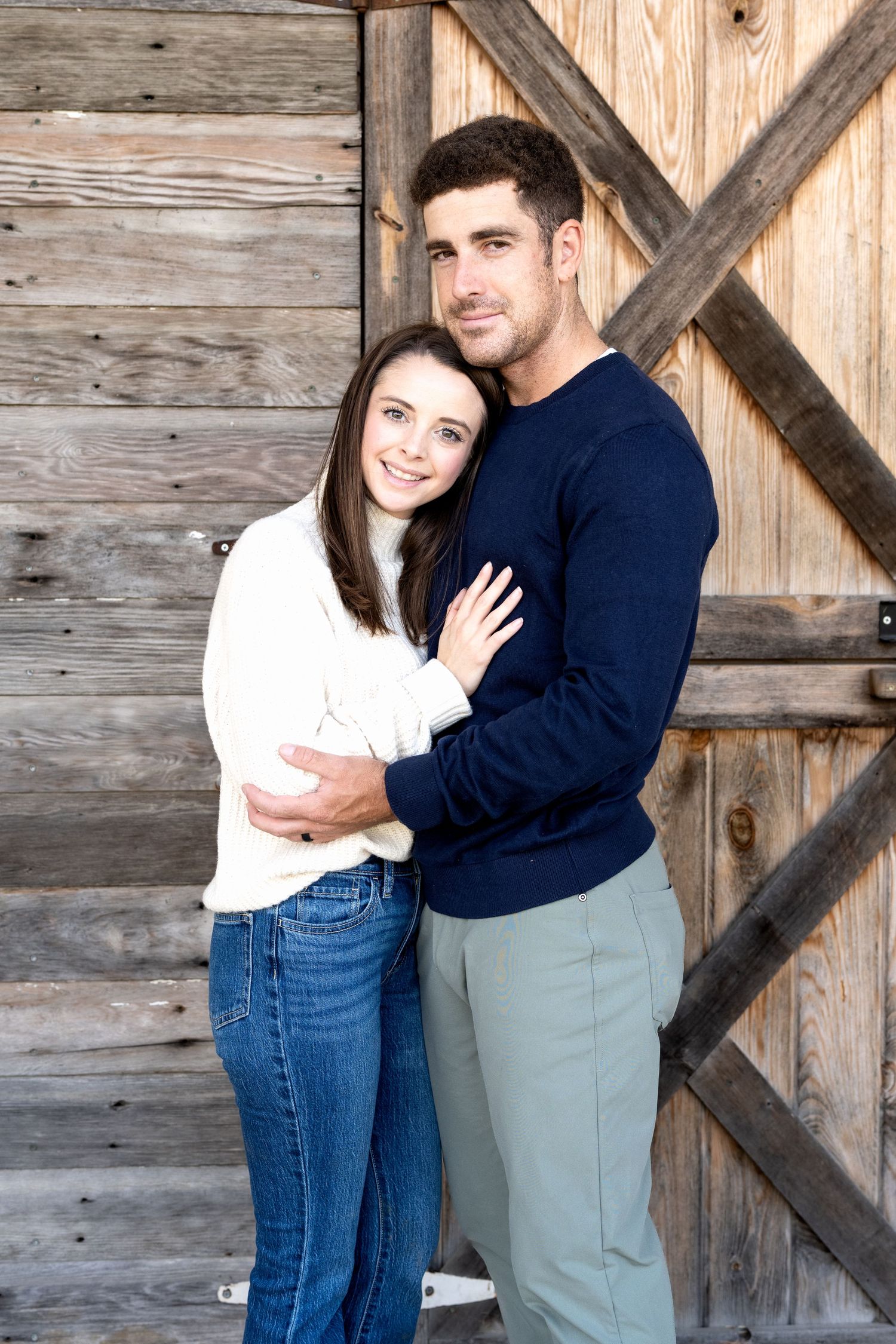 A young couple embraces warmly in front of a rustic wooden barn door, wearing casual sweaters and jeans.