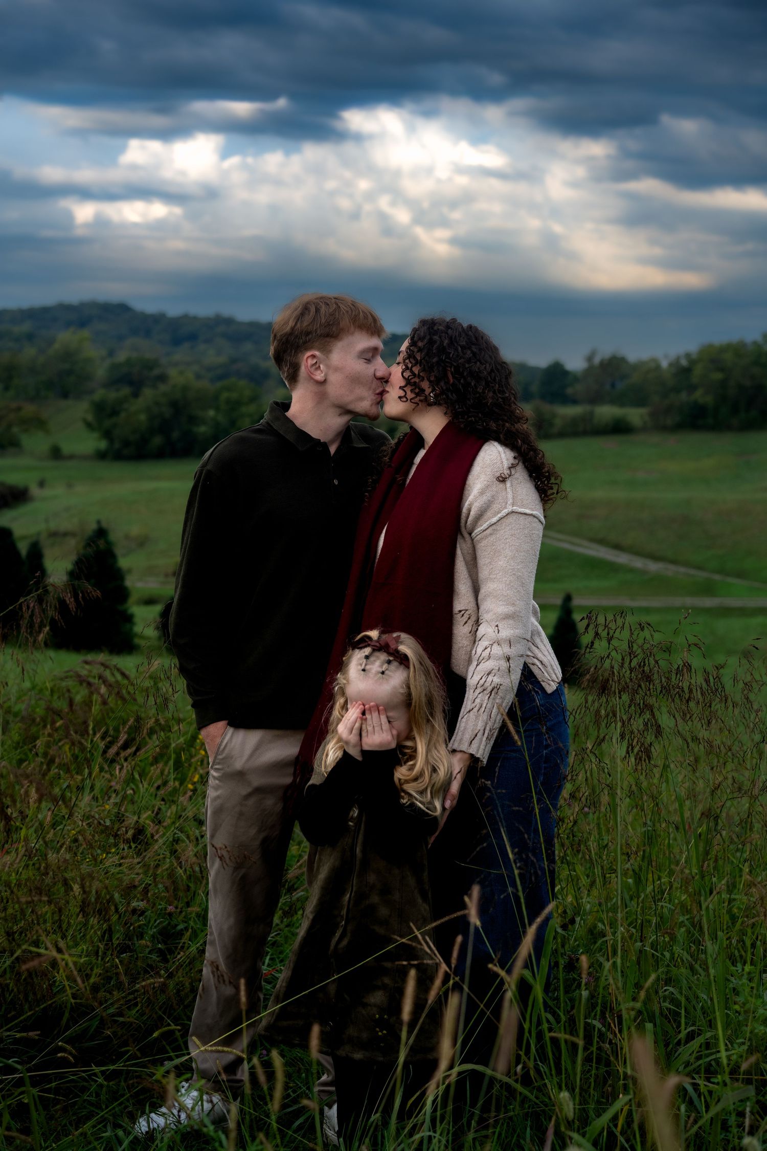 A couple shares a kiss in a grassy hillside field under a dramatic cloudy sky while a young girl covers her face in front of them.