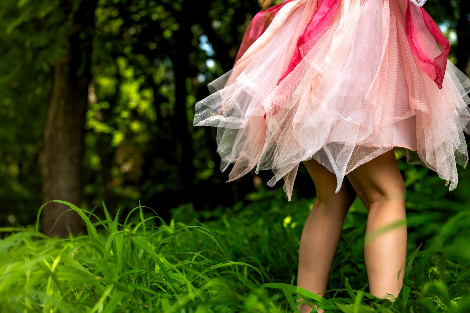 Young girl in pink tutu dress walking barefoot through lush green grass in a forest setting.