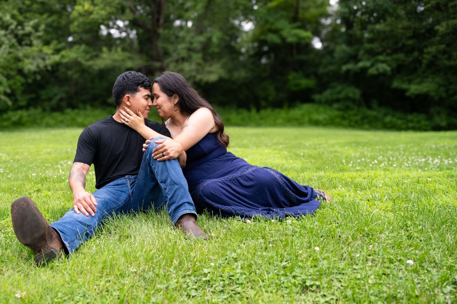 lifestyle portrait of a portrait of A couple sits on green grass in a park, leaning in for a kiss, wearing casual blue and black outfits.