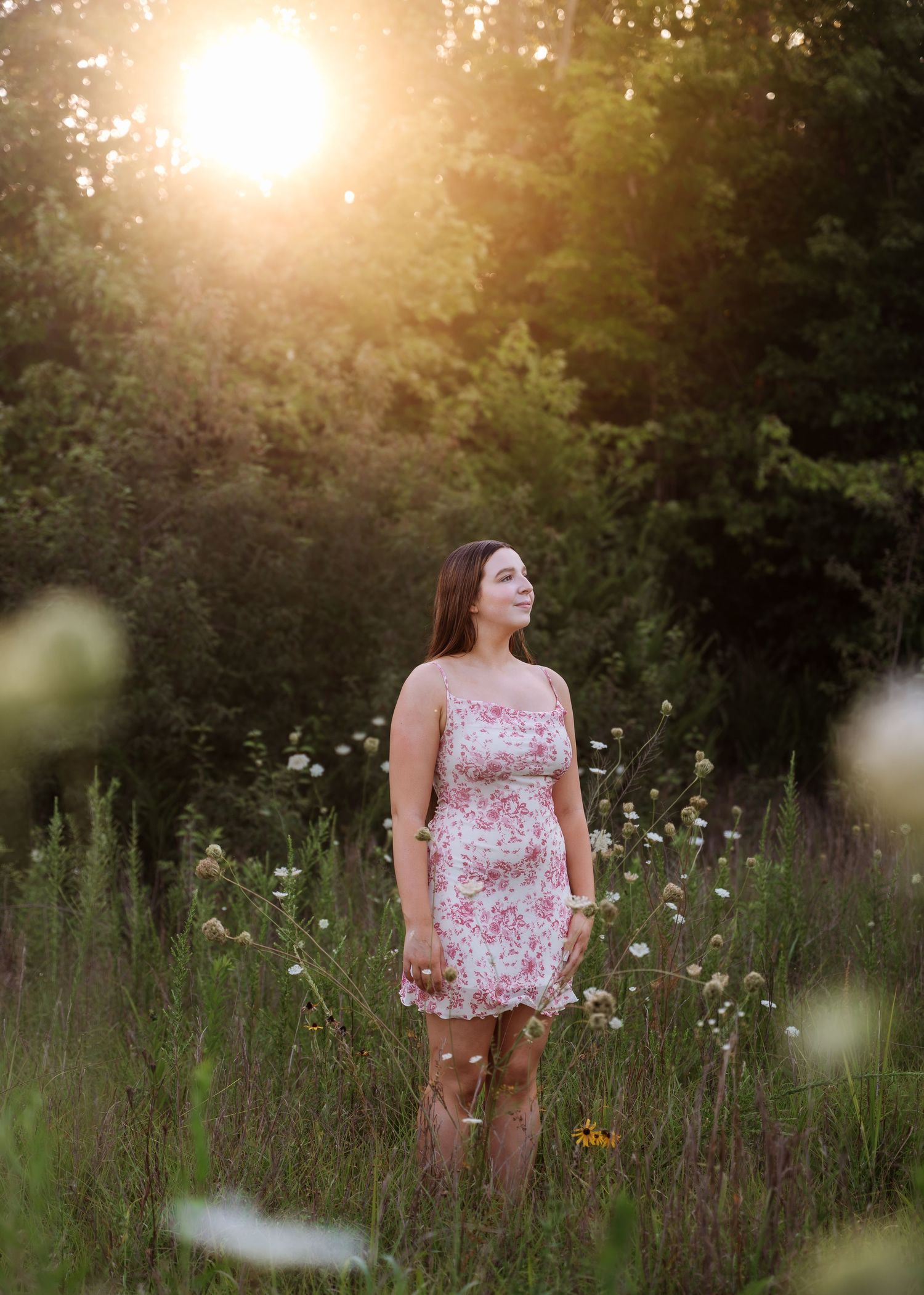 lifestyle portrait Young woman in floral dress standing in wildflower meadow bathed in golden sunset light with lush green trees behind her.