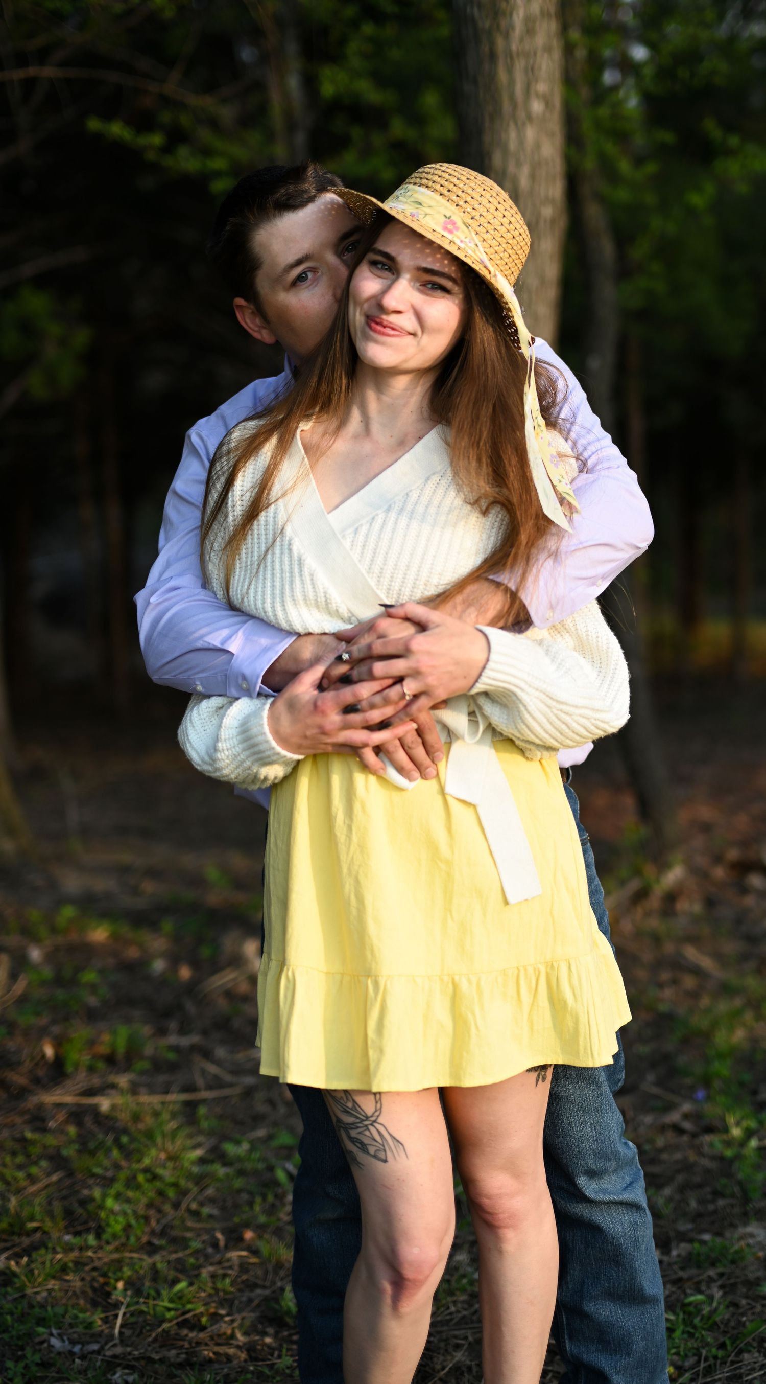 A man embraces a smiling woman in a yellow dress and straw hat from behind in a forest setting.