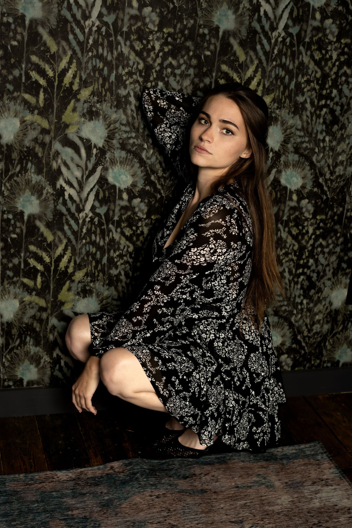 Young woman in floral black dress poses gracefully against dark botanical wallpaper on hardwood floor.