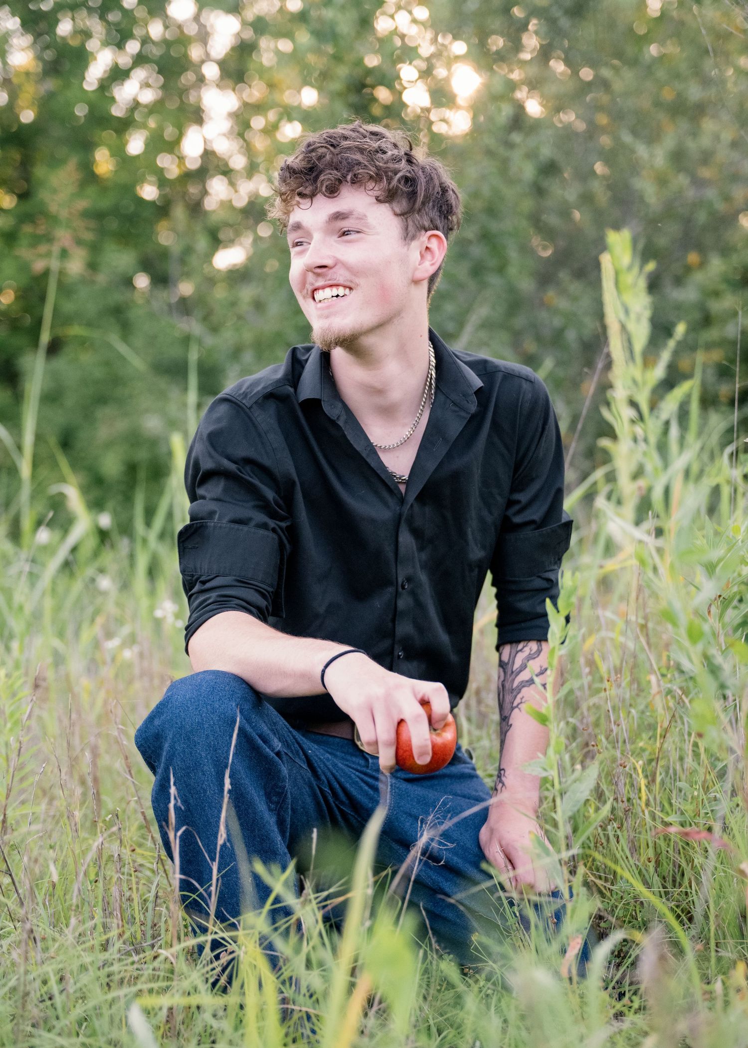 portrait of a Laughing young man with tattoo crouching in tall grass holding a red apple outdoors.