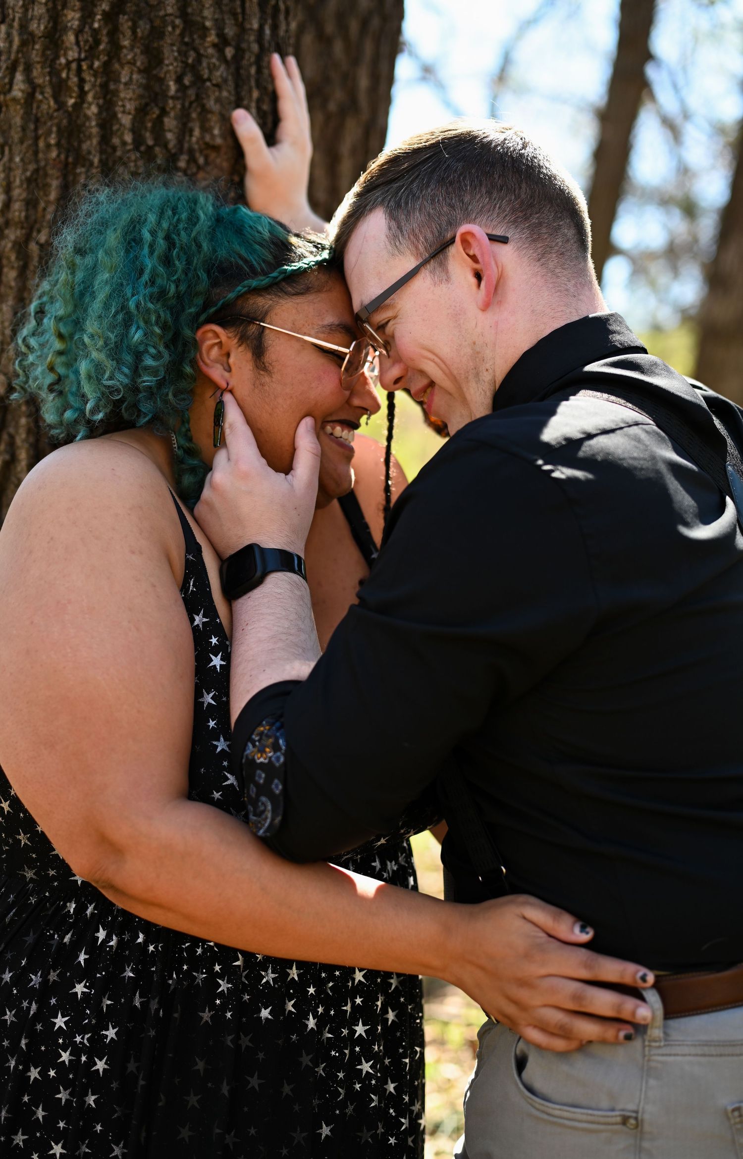 Interracial couple smiling and embracing closely against a tree trunk outdoors.