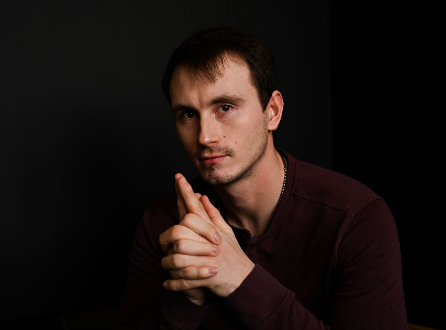 A young man in a maroon shirt clasps his hands together, posing against a dark studio background.