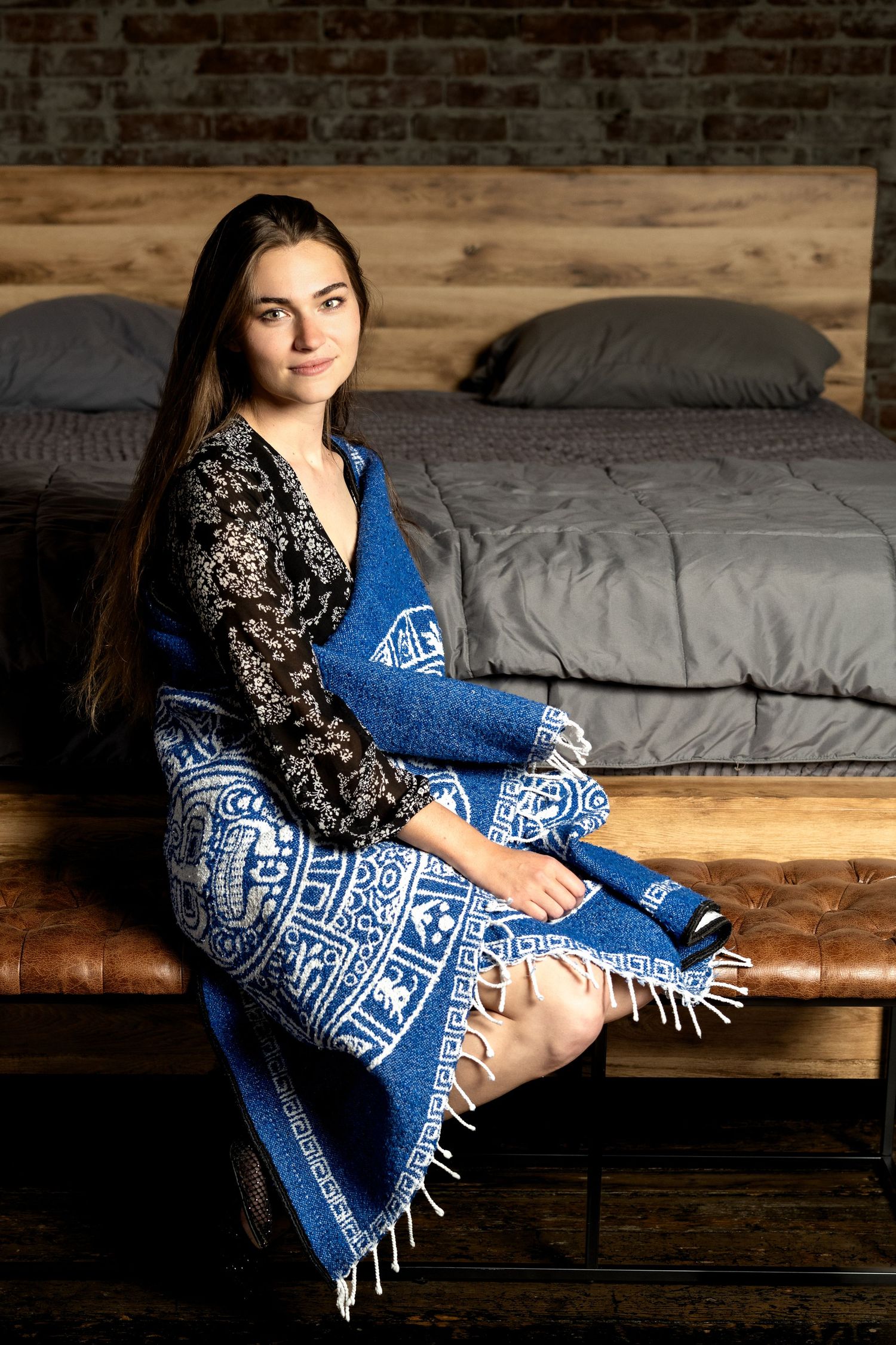 Young woman with long dark hair sits on a bed frame wrapped in a blue and white patterned blanket with fringe.