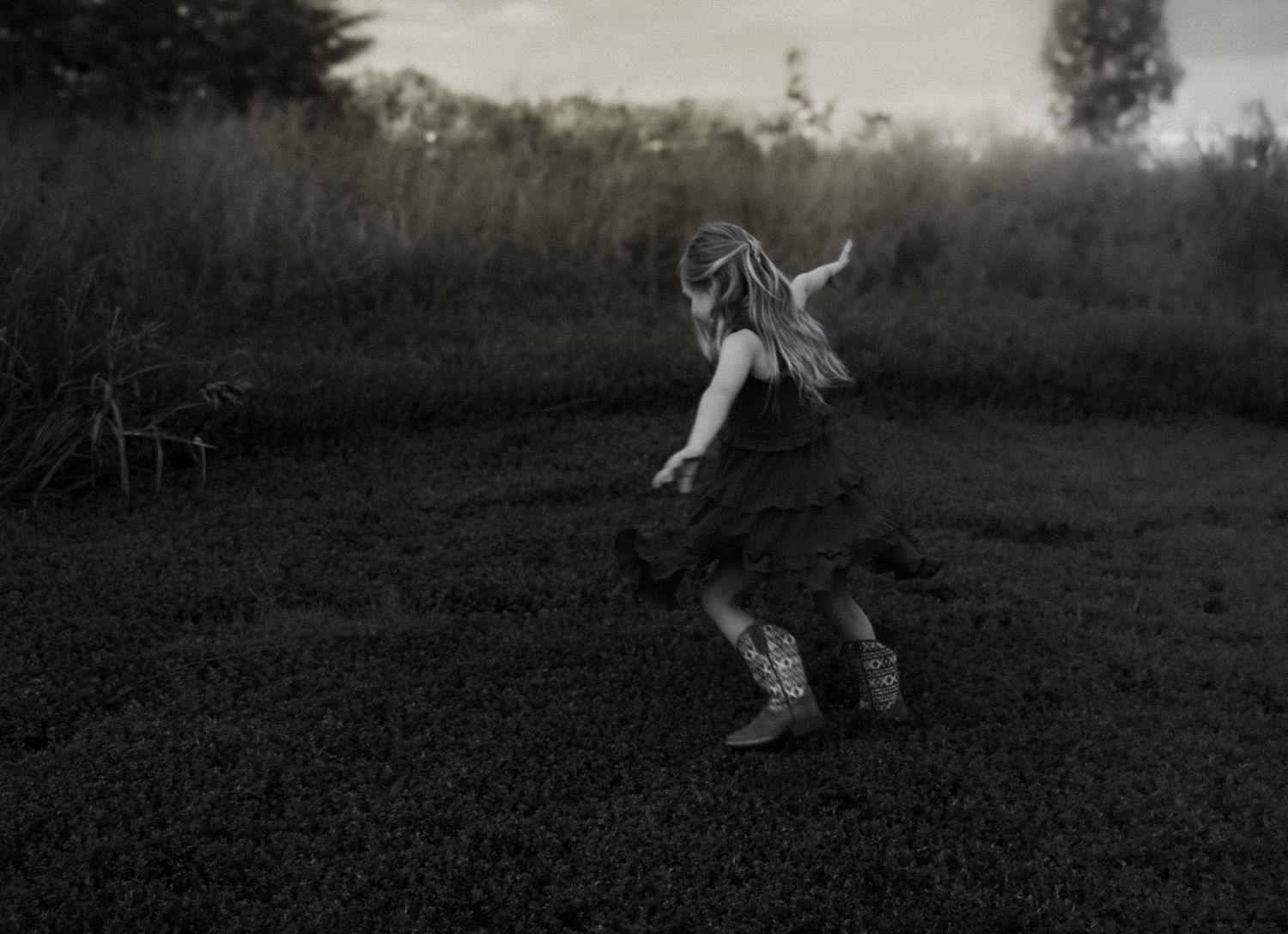 A young girl in a dark dress and boots runs through a moody, overgrown field in a black and white photo faceless family photography