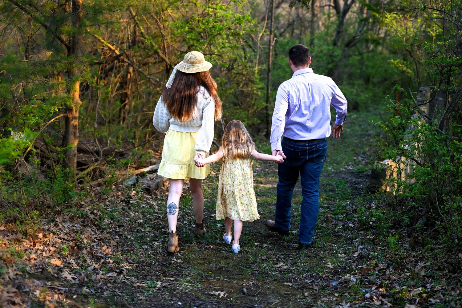 Family of three walking hand-in-hand down a forest path with a dog, mother in yellow dress and hat.