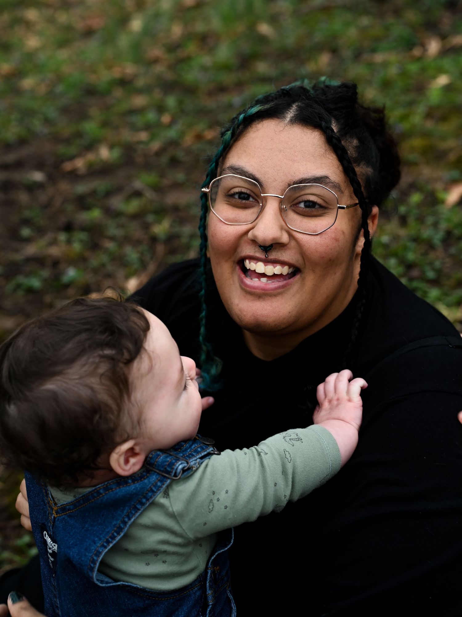 Smiling mother with teal braids and glasses holding laughing baby in denim overalls outdoors.