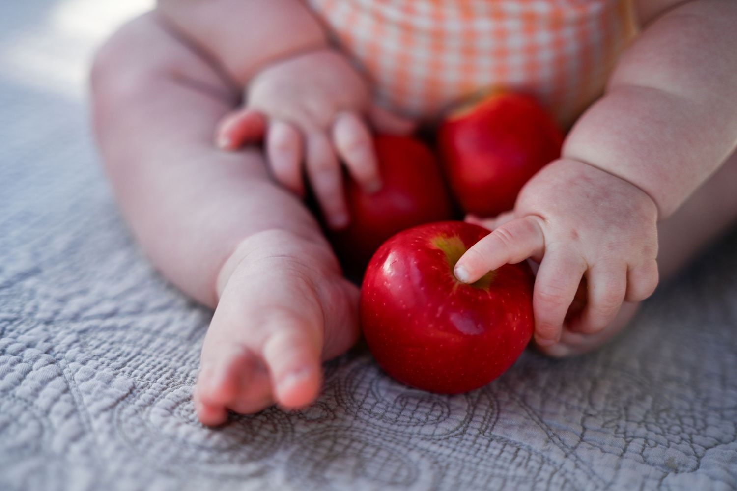 A baby's chubby hands and feet surrounded by three red apples on a gray blanket.