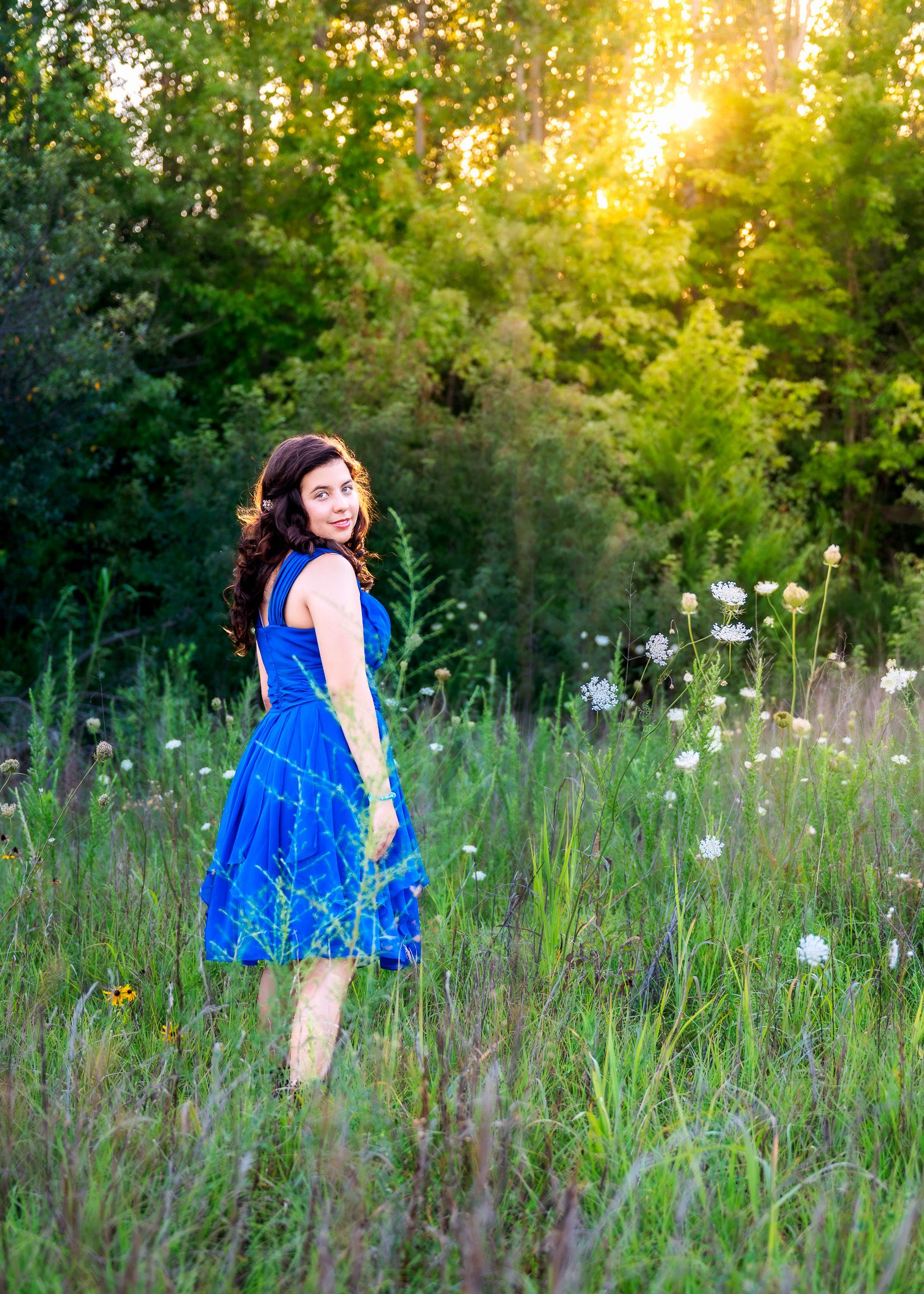 Young woman in blue dress standing in sunlit meadow with wildflowers and lush green trees in background.