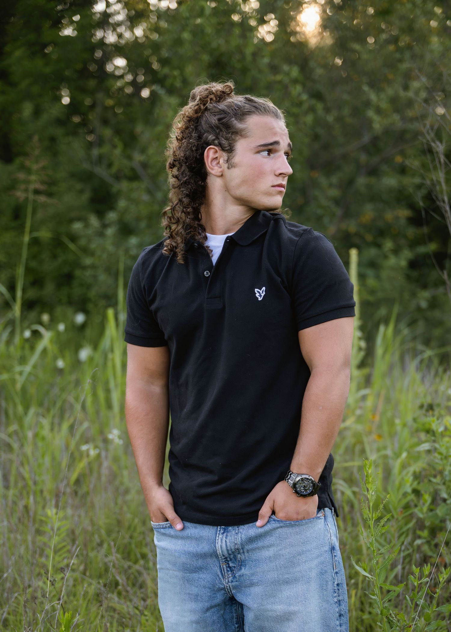 portrait of a Athletic young man with ponytail wearing black polo stands in green field.