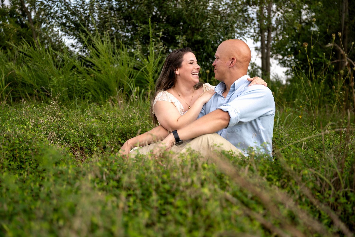 lifestyle portrait of a Couple sitting in lush green grass, smiling and embracing each other outdoors surrounded by trees.
