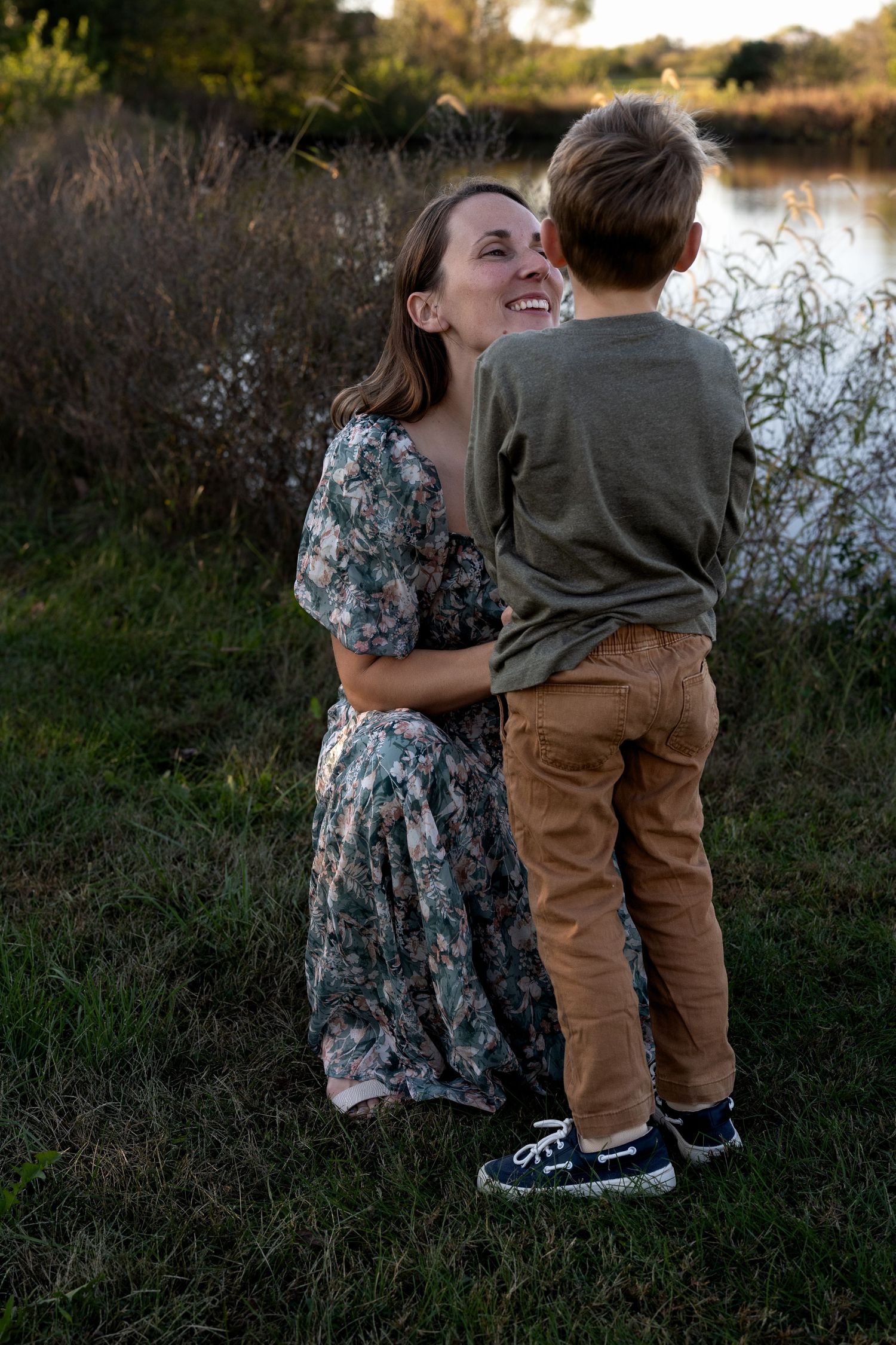 Smiling woman in a floral dress kneels on grass by a pond, hugging a young boy in a green shirt, tan pants and navy sneakers.