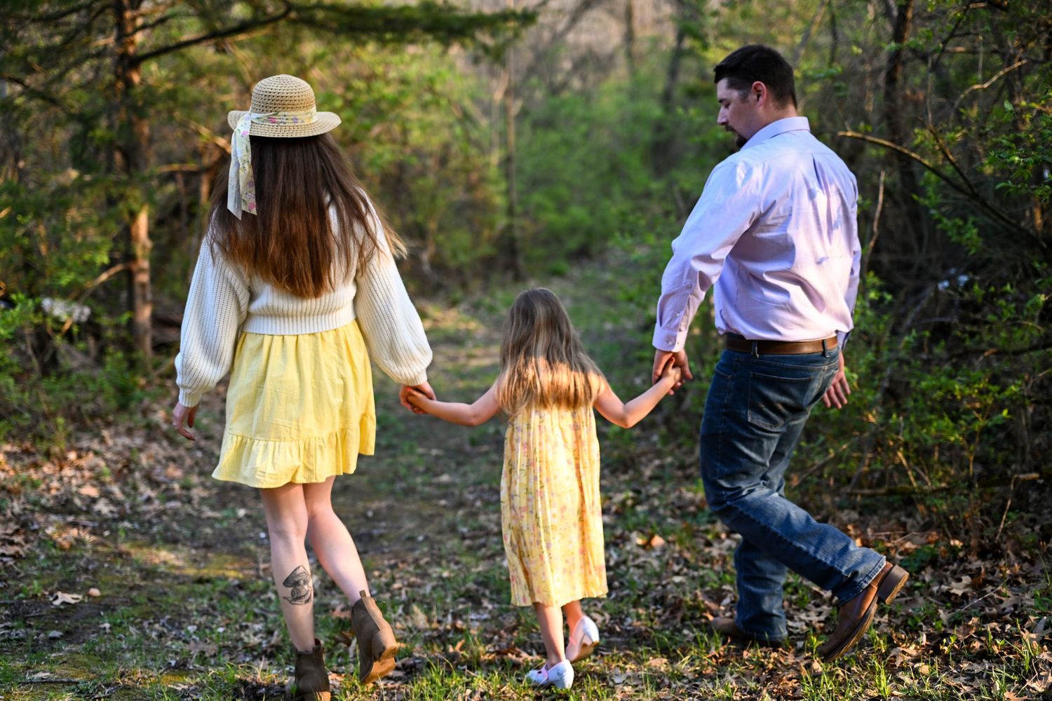 A family of three walks hand-in-hand along a wooded path, with parents and a young girl in matching yellow dress.