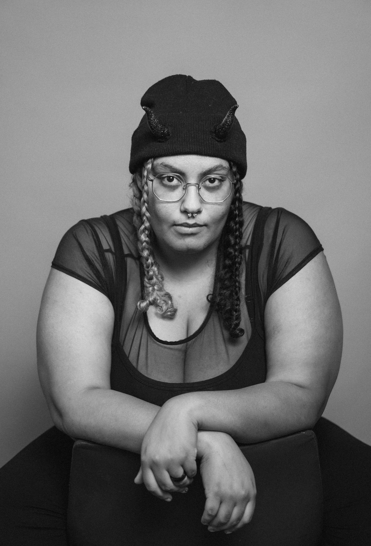 Black and white portrait of a person wearing a beanie, braids, and glasses, leaning forward with arms crossed.