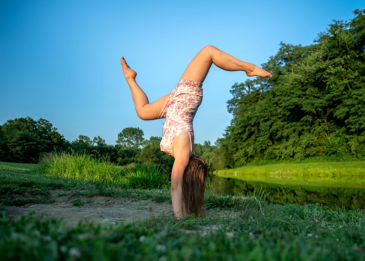 lifestyle portrait of a Young girl performing a handstand on grass near a pond surrounded by lush green trees under a clear blue sky.