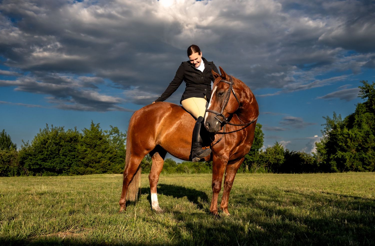 lifestyle portrait of a Woman in equestrian attire riding a chestnut horse on green field under dramatic cloudy sky.
