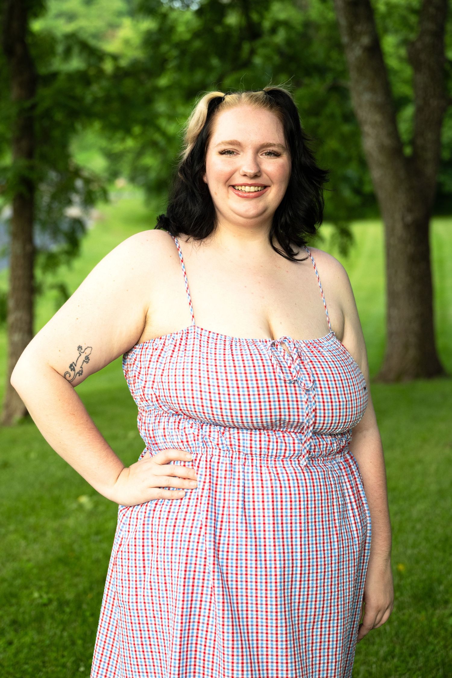 Smiling young woman with tattoo wearing a red and white checkered sundress posing outdoors in a green park.