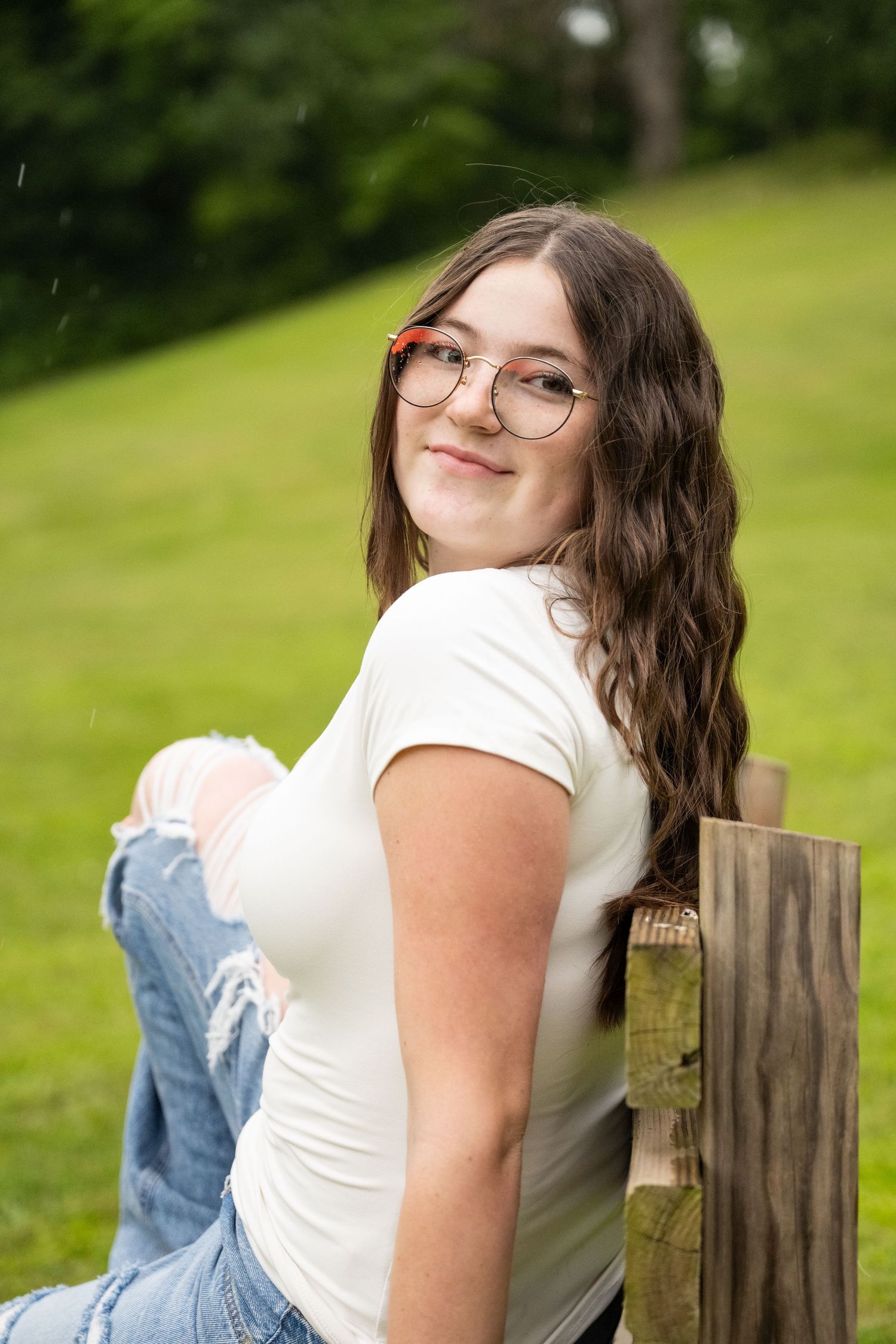 portrait of a Teen girl in white tee and ripped jeans sitting on wooden bench smiling in green park.