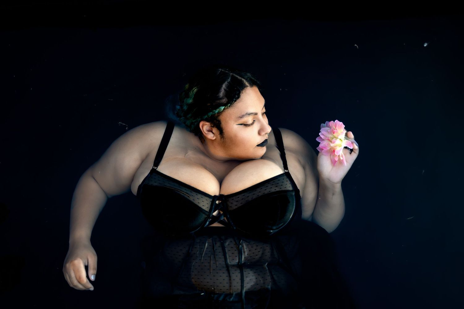Plus-size woman in black lingerie corset holds a pink flower against a dark background.
