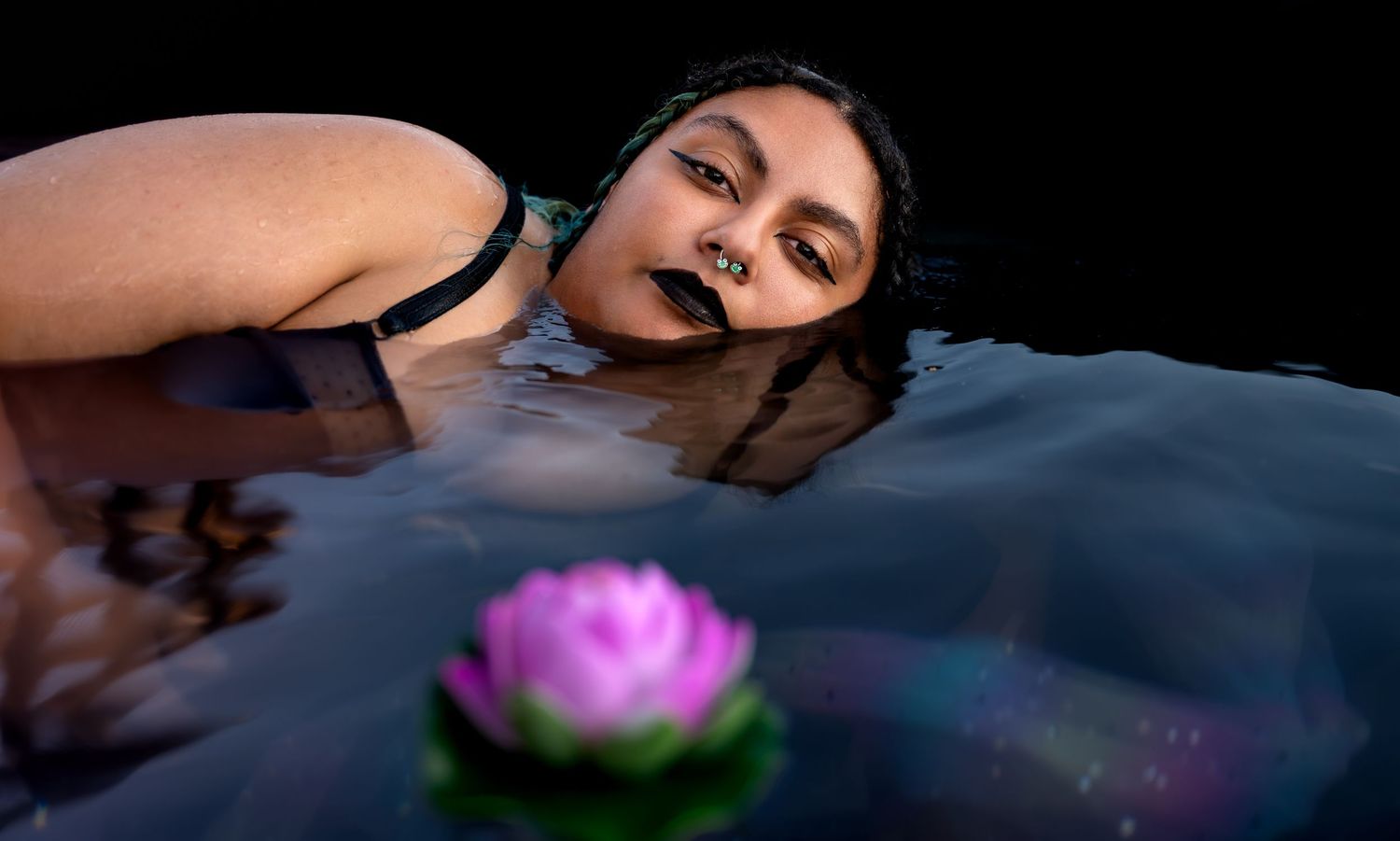 Woman with dark makeup floating in water beside a pink lotus flower at night.