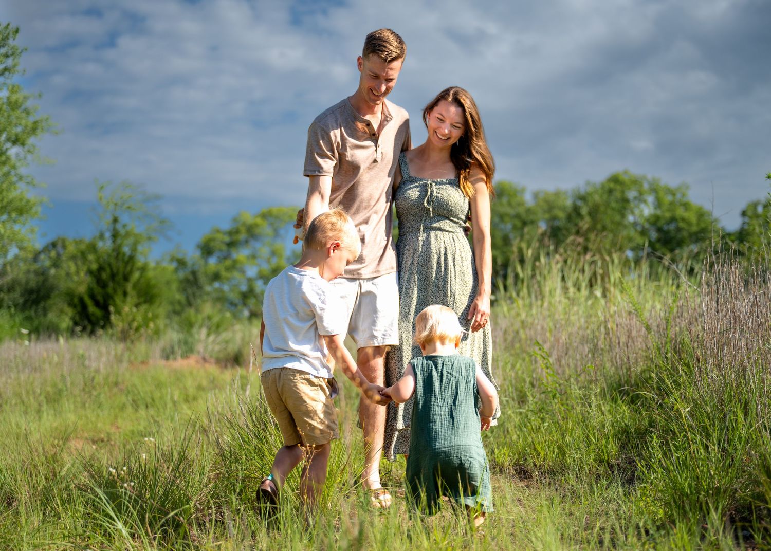 Family of four enjoying a sunny day in a lush green meadow, parents smiling with two young children.