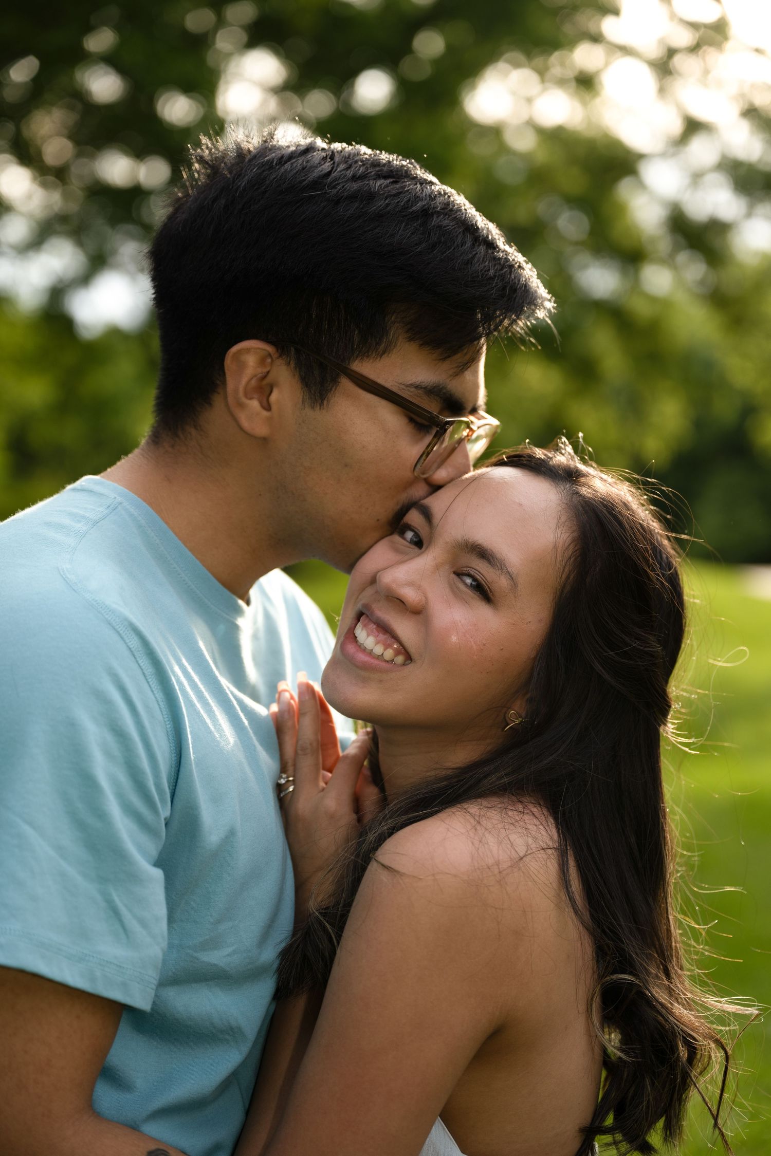 portrait of a Woman smiles brightly as man kisses her cheek in a warm golden sunlit park setting.