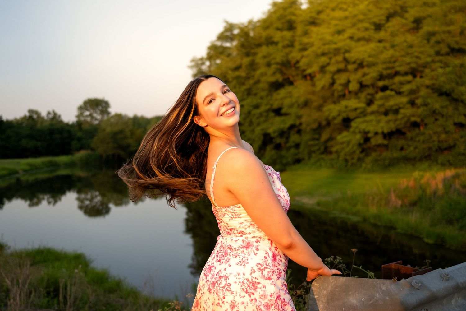 Young woman in floral dress smiling joyfully by a serene pond, hair flowing, surrounded by lush green trees at sunset.