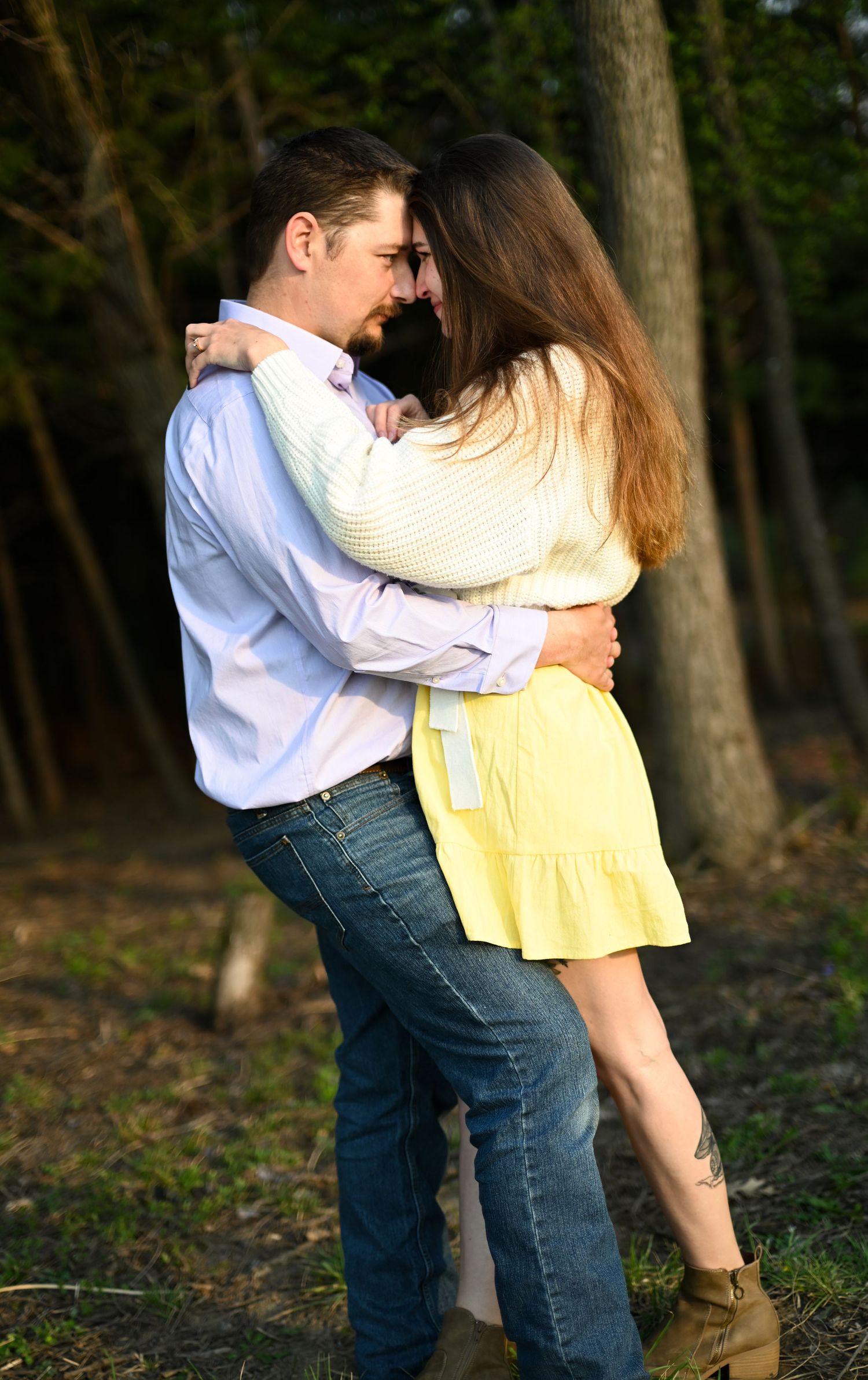 A romantic couple embraces and almost kisses outdoors in a sunlit forest setting.