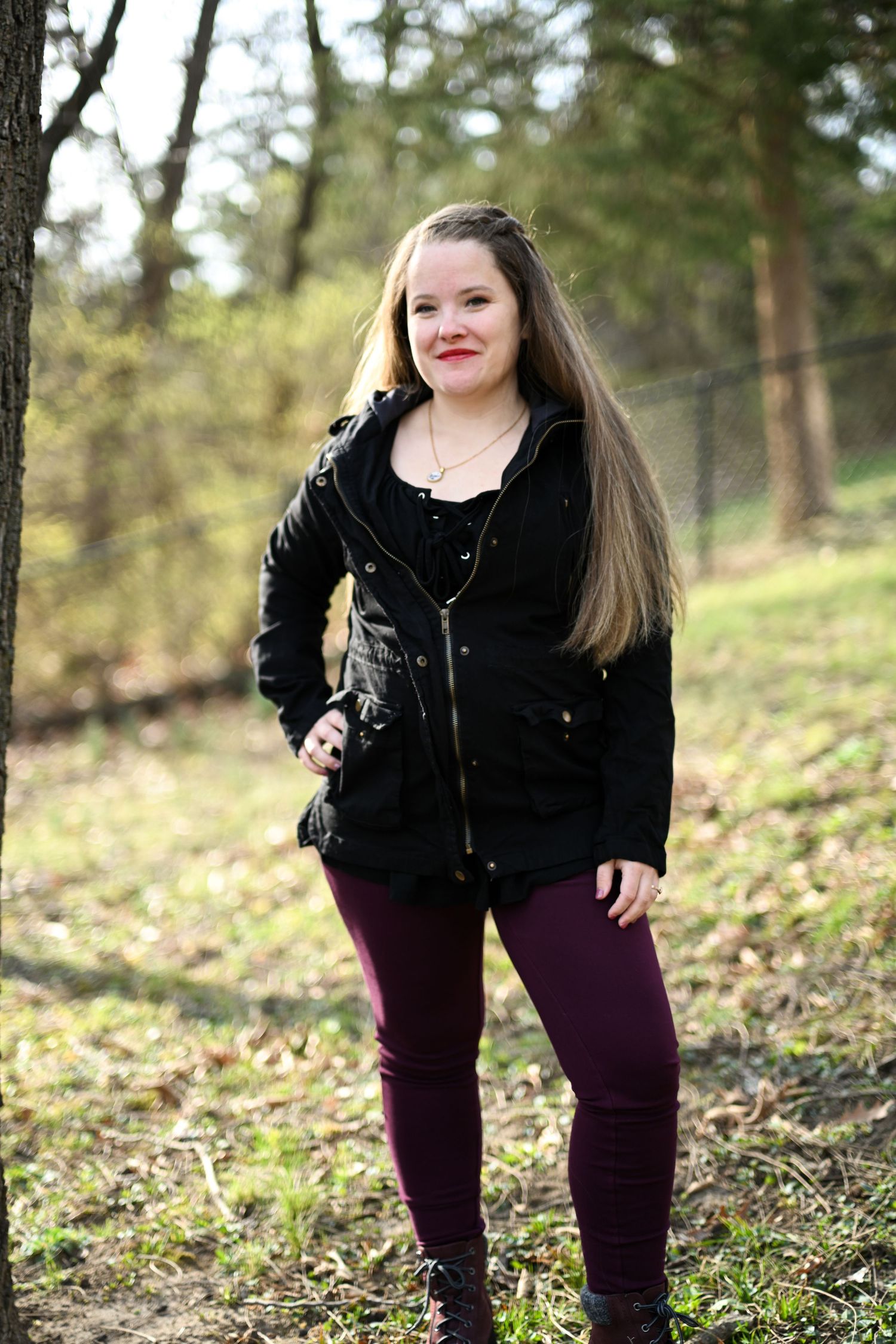 Young woman in black jacket and purple jeans smiling while standing in forest.