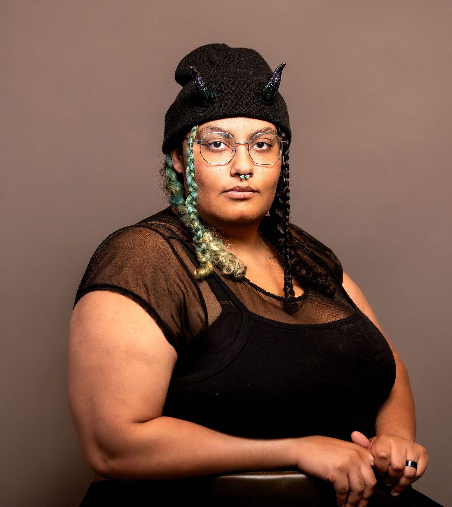 Person wearing black sheer top, black hat with horns, glasses, and teal braids, posing against a gray background.