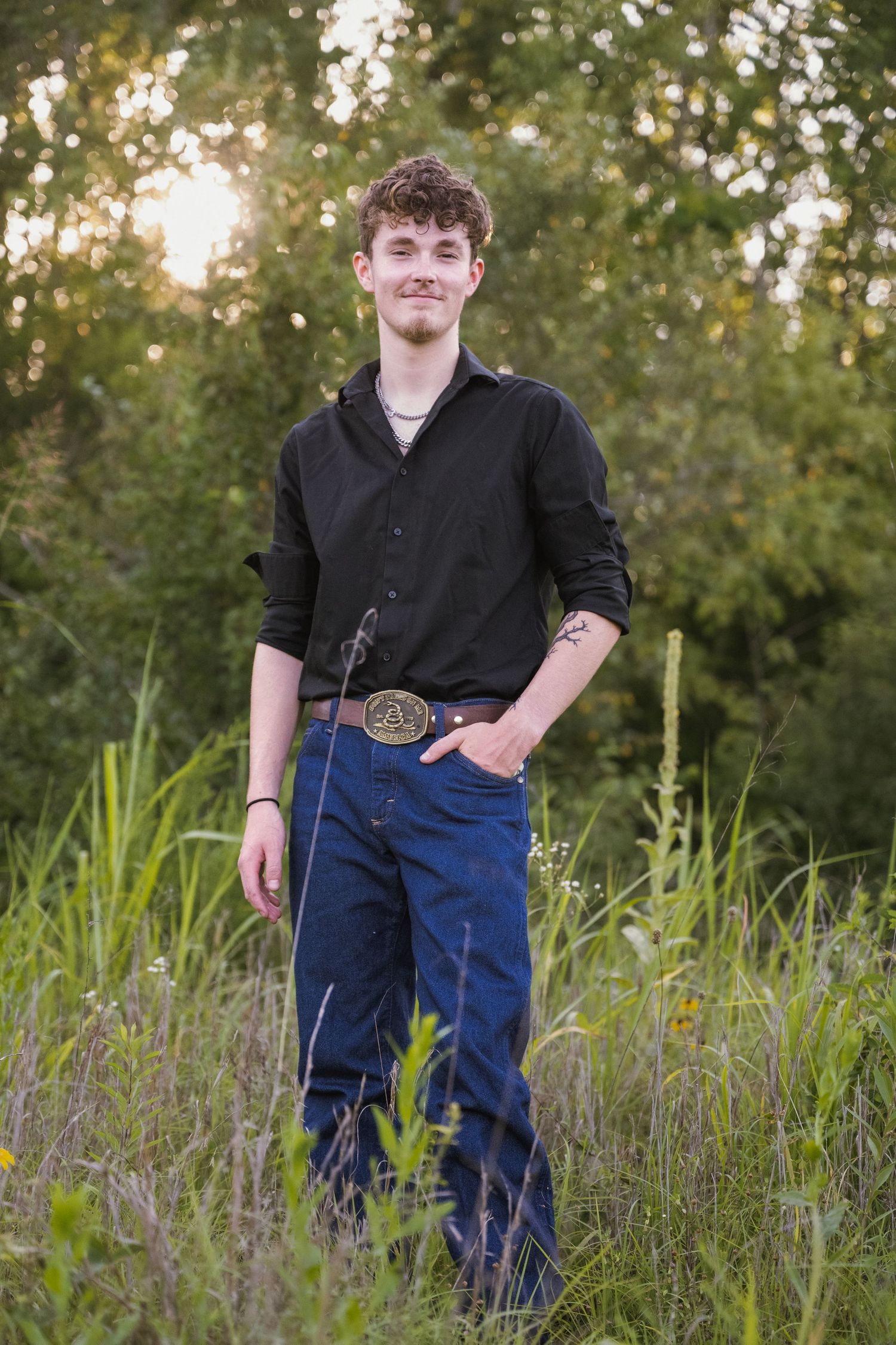 portrait of a Smiling young man in black shirt and jeans with decorative belt buckle standing in tall grass.