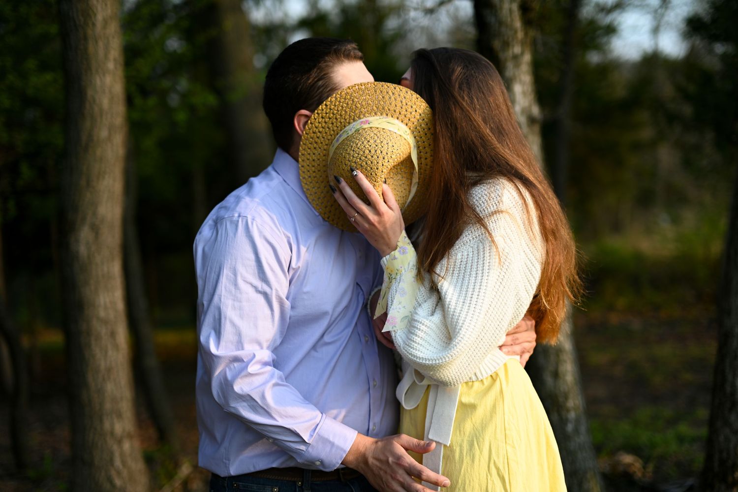 A couple shares a kiss hidden behind a straw hat outdoors in a sunny wooded setting.