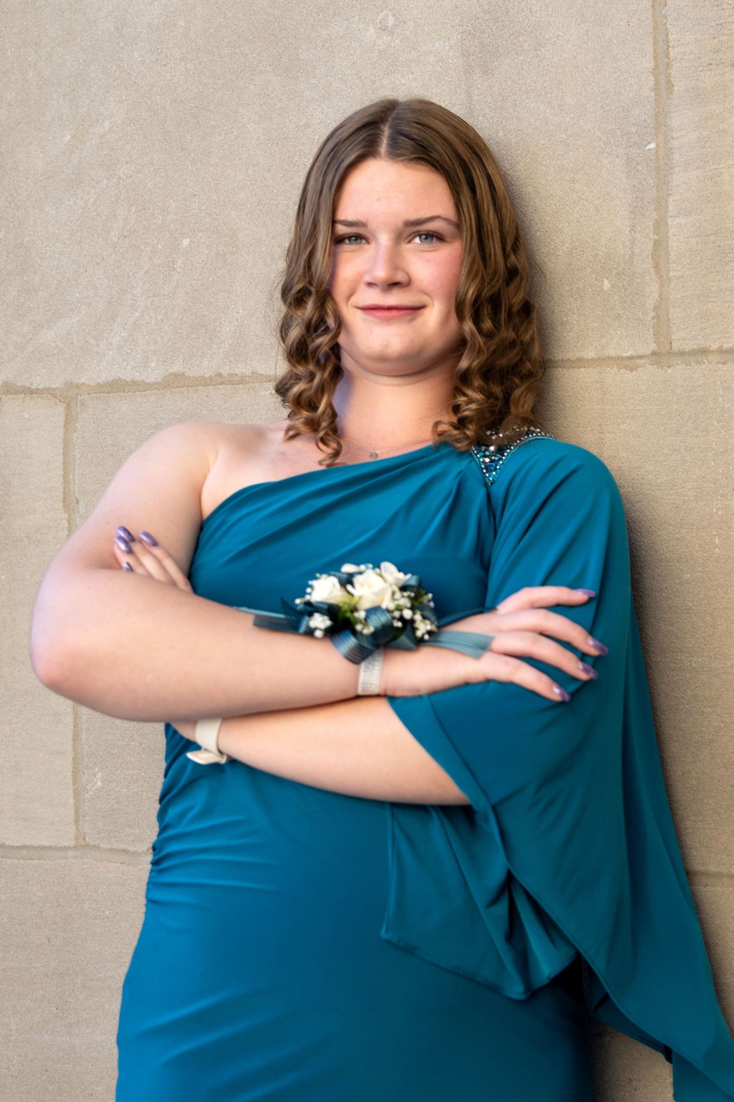 Young woman in teal one-shoulder gown with wrist corsage poses arms crossed against stone wall.