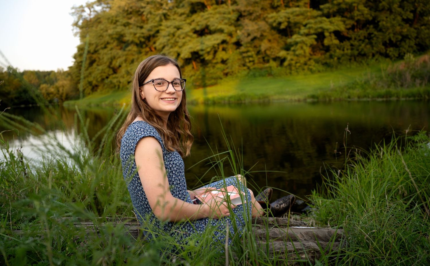 lifestyle portrait of Young woman with glasses sitting by a serene pond surrounded by lush green grass and trees in nature.