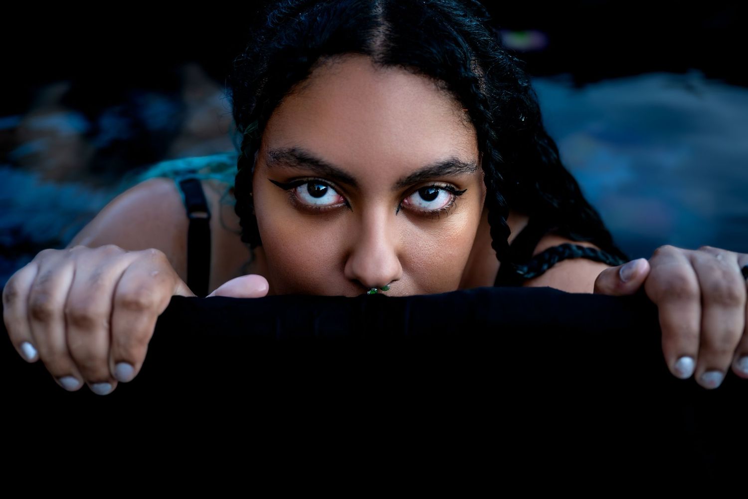Young woman with dark hair and dramatic eye makeup peering over a dark surface with an intense gaze.