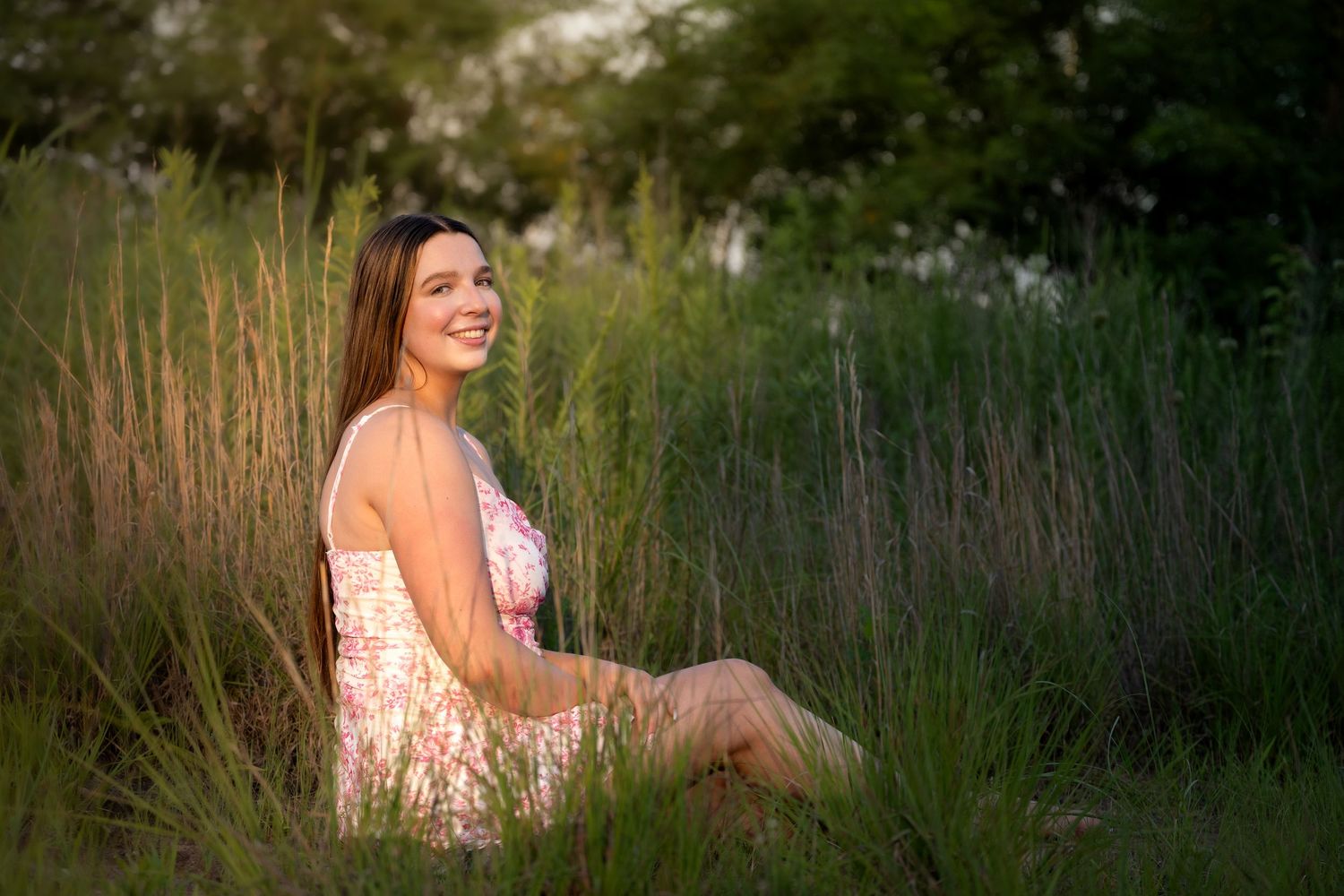 lifestyle portrait of a Young woman in floral dress sitting in tall green grass, smiling outdoors in warm golden sunlight.