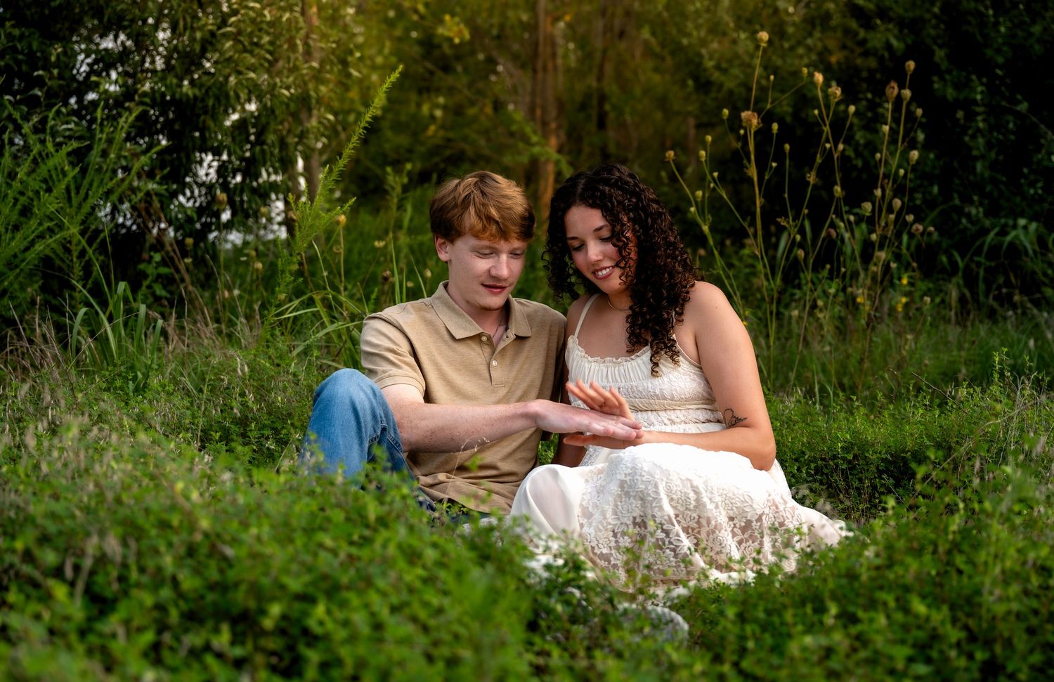 lifestyle portrait of a young couple sitting together in lush green meadow, sharing a tender moment outdoors in natural setting.