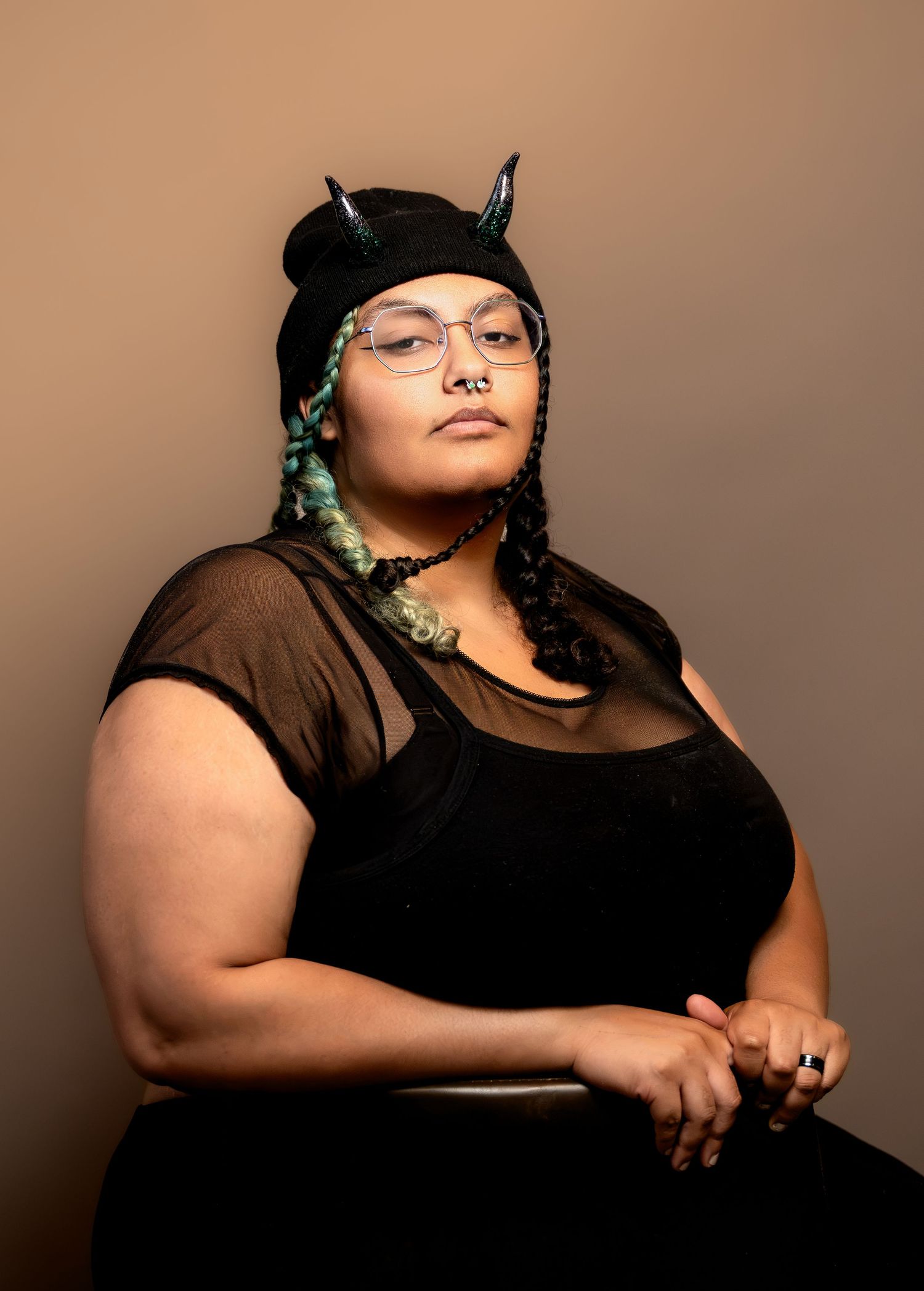 Person wearing black beanie with horns, glasses, and braids poses confidently in a black outfit against a brown background.