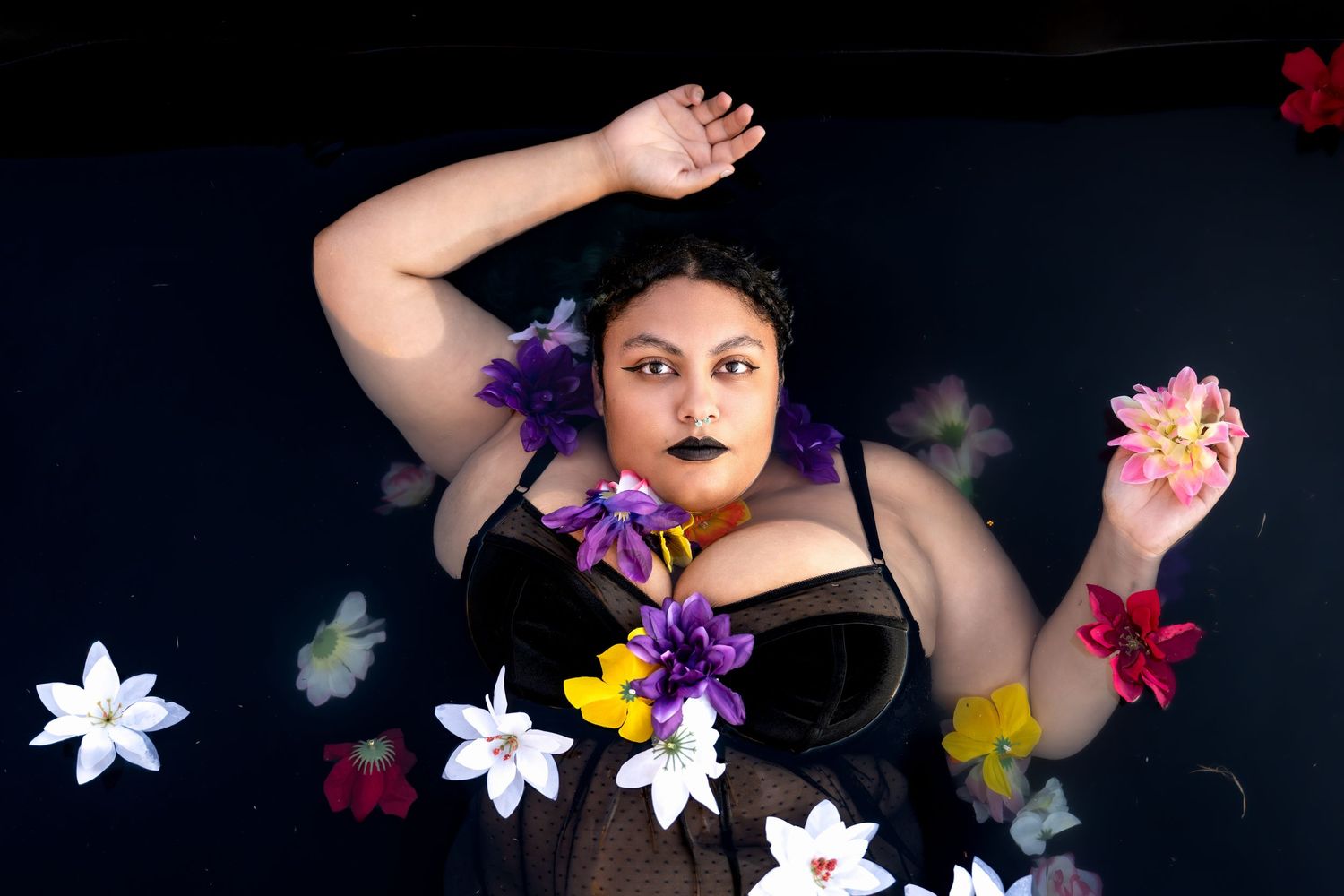 Woman in black corset surrounded by colorful flowers floating in dark water, striking black lipstick.