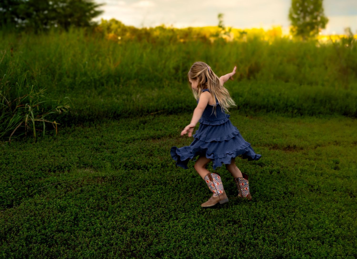 A young girl in a blue ruffled dress and cowboy boots twirls joyfully in a lush green meadow at sunset. Faceless family photography