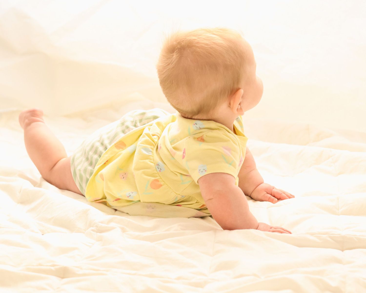 Baby in yellow floral romper doing tummy time on a soft white blanket.